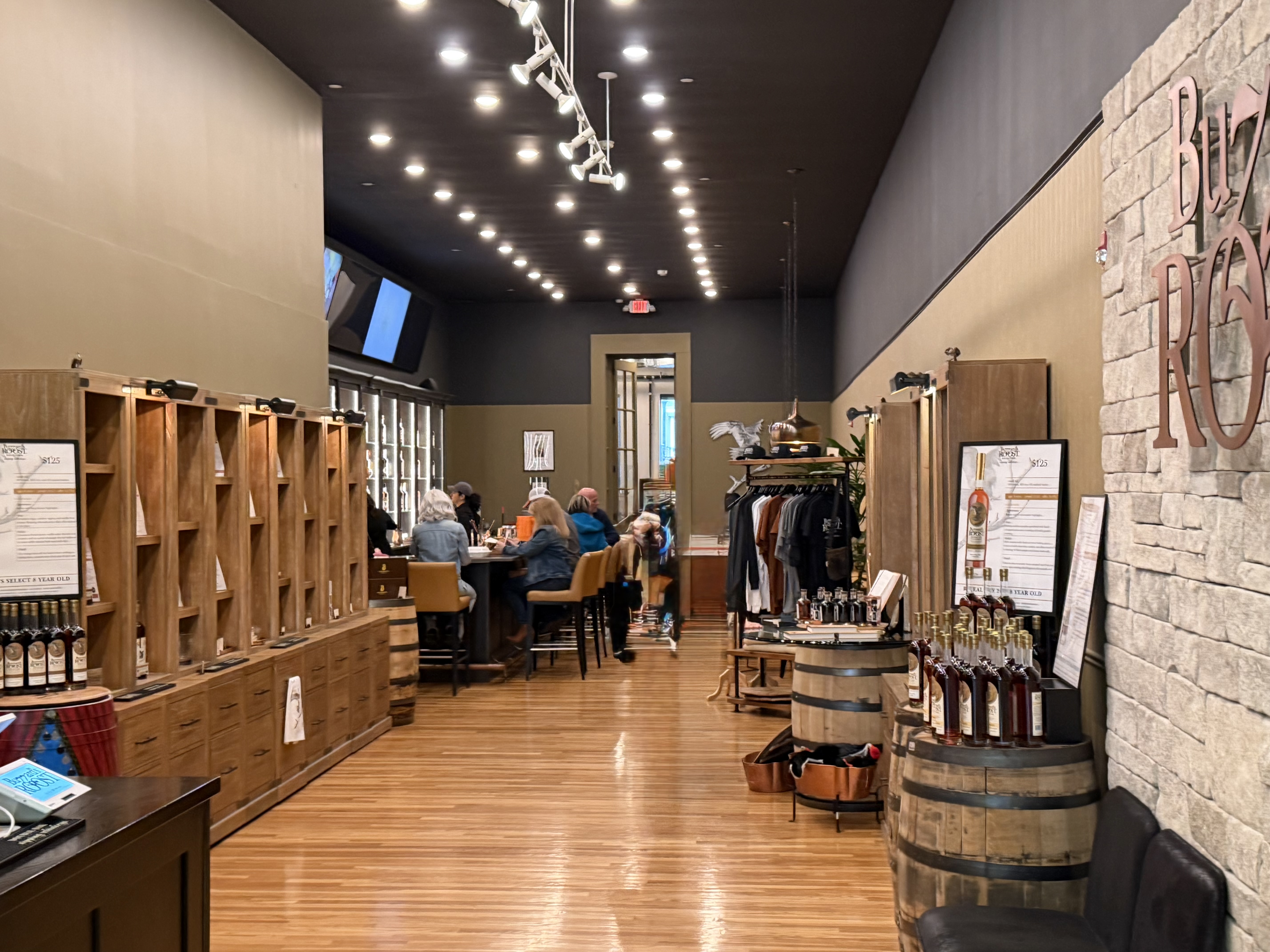 Buzzard's Roost tasting room interior with bottle shelving, seated guests, barrel displays, and the Buzzard's Roost logo on brick wall