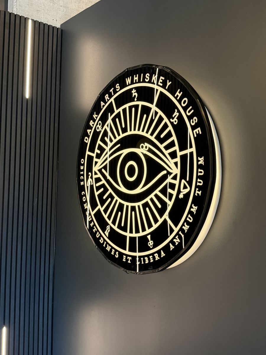 Dark Arts Whiskey House emblem, a gold eye inside a circular dial on a black wall