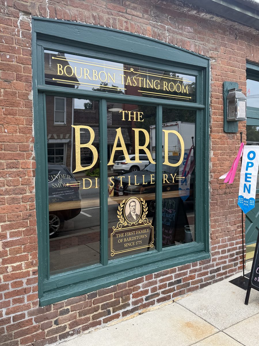Window lettering reading The Bard Distillery Bourbon Tasting Room with the First Family of Bardstown crest